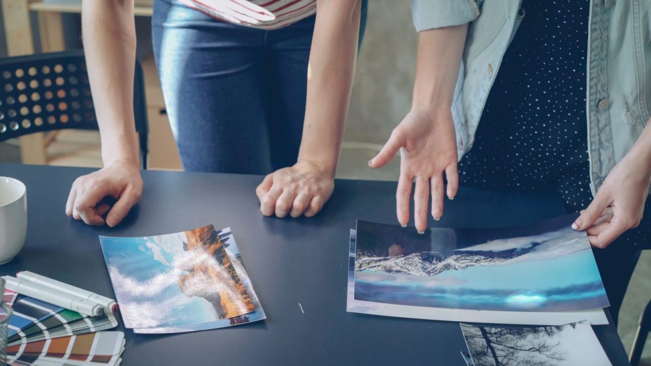 Two people reviewing printed landscape photographs spread out on a table indoors