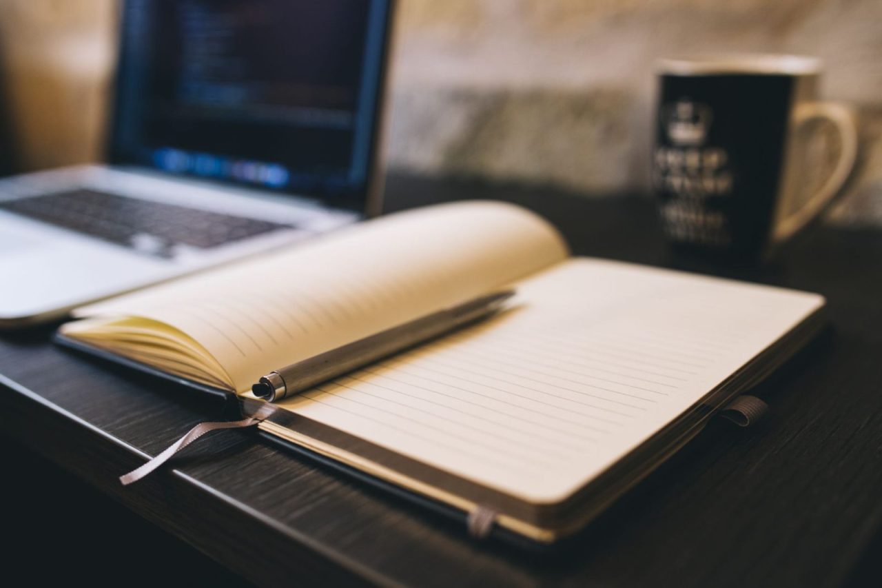 Open notebook with pen on a wooden desk next to a laptop, suitable for blogging or work themes