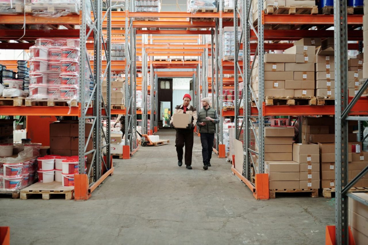 deux employés manipulent un colis dans un entrepôt avec des étagères remplies de cartons et produits