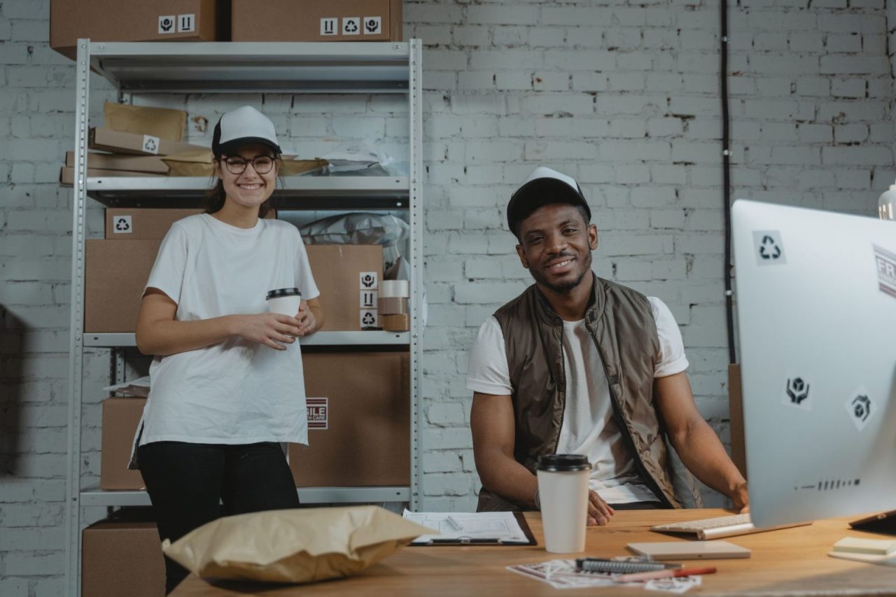 Two warehouse employees in casual attire smiling while handling packages in a courier setting